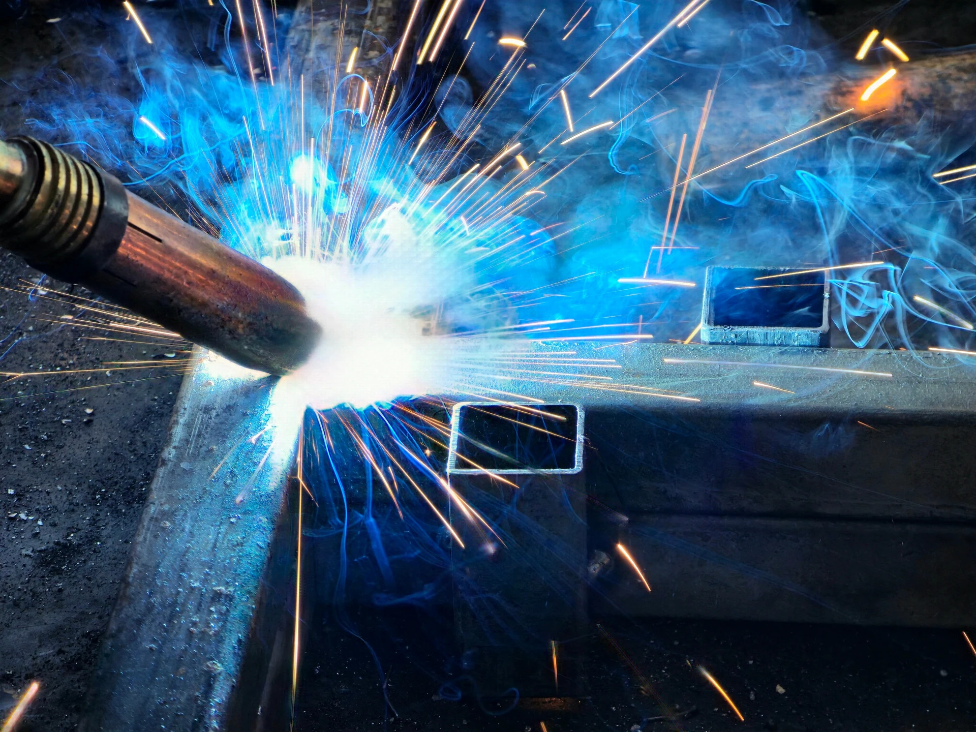 Soudeur professionnel travaillant dans un atelier industriel sombre, portant un masque de protection automatique et des gants de sécurité, effectuant une soudure à l'arc électrique avec production d'étincelles dorées et fumées colorées bleu-violet
